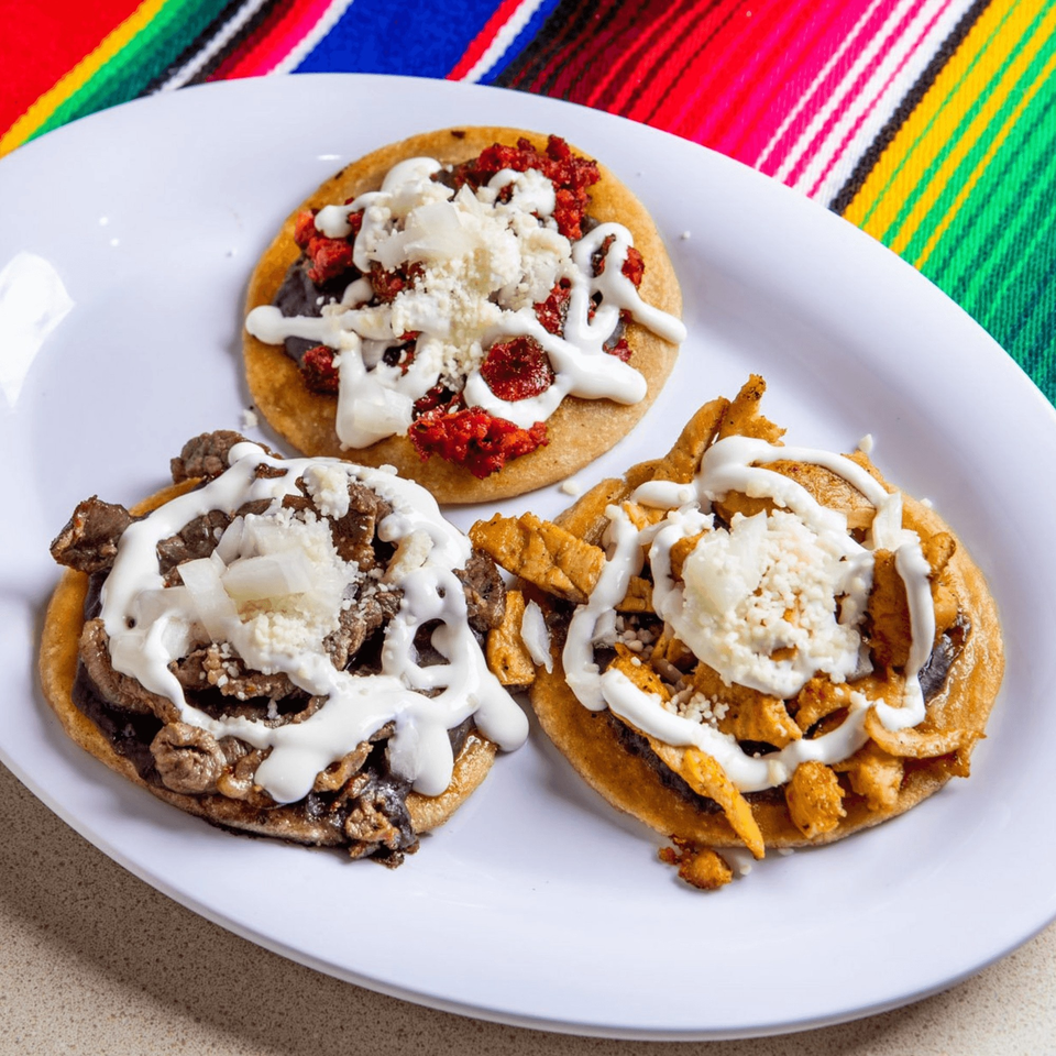 Three Homemade Taqueria sopes with various toppings, crema, and cheese on a white plate.
