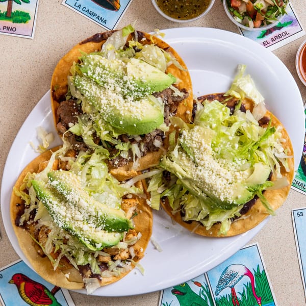 Three Homemade Taqueria tostadas with meat, lettuce, avocado, and cheese, surrounded by Loteria cards.