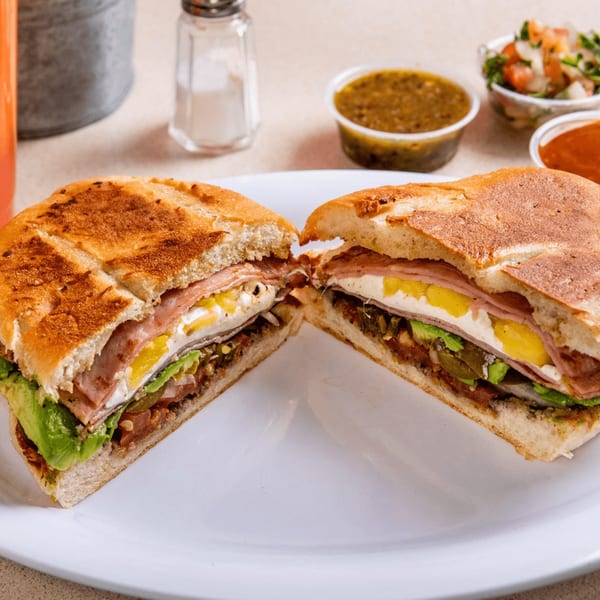 A large Mexican torta sandwich, cut in half, revealing layers of meat, cheese, avocado, and vegetables.